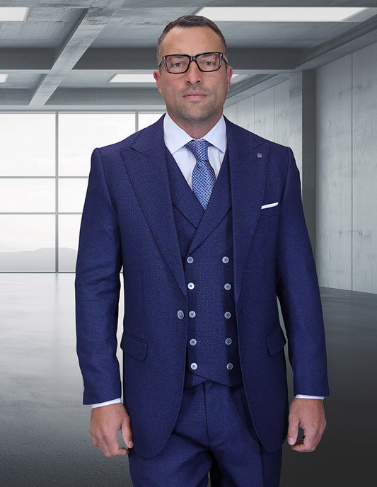A man in a STATEMENT CLOTHING | NEIVE-SAPPHIRE suit by Statement Clothing, with a light blue shirt, patterned tie, and glasses, stands in a modern empty room with large windows and concrete walls.