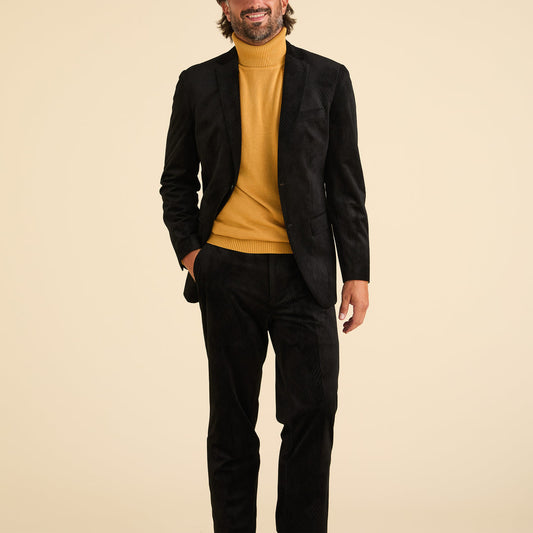 A man smiles in front of a beige background, wearing the Inserch Velvet Diagonal Suit BL275-01 Black by Inserch with paisley print lining and modern fit pants, paired with a mustard yellow turtleneck sweater.