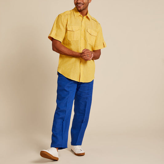 A person in a yellow short-sleeve shirt, Inserch Linen Pants P3116-00190 Estate Blue, and white sneakers stands against a plain background, smiling with hands clasped.