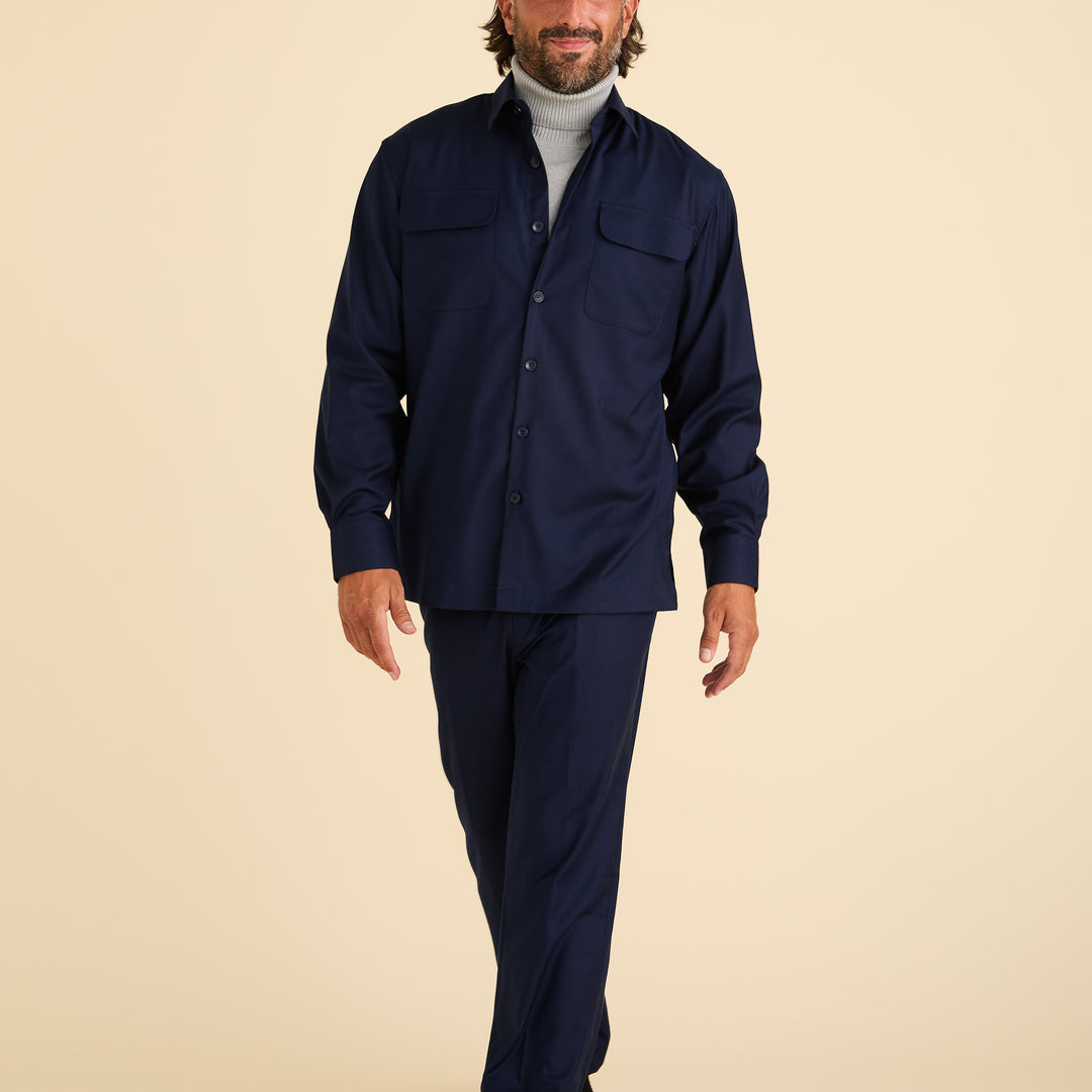 A man walks against a beige background, wearing Inserch Modern Fit Pants P3199S-36 Ink and a navy blue button-up shirt layered over a light grey turtleneck.