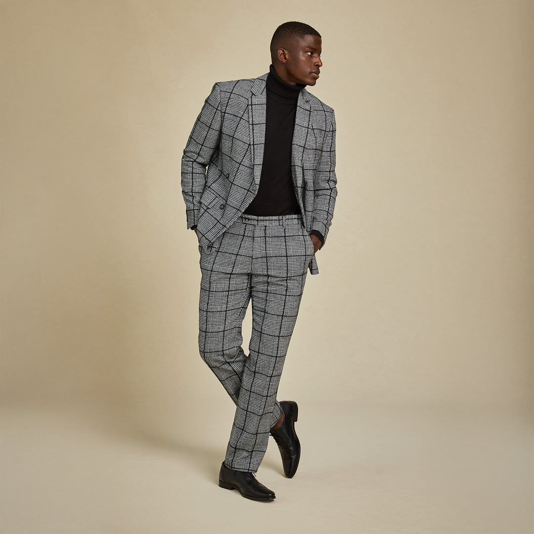 A man wearing Inserch Glenn Check Pants P504-41 Black/White stands against a beige background, paired with a black turtleneck and dress shoes, looking to the side with one leg crossed over the other.