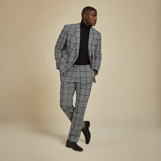 A man wearing Inserch Glenn Check Pants P504-41 Black/White stands against a beige background, paired with a black turtleneck and dress shoes, looking to the side with one leg crossed over the other.