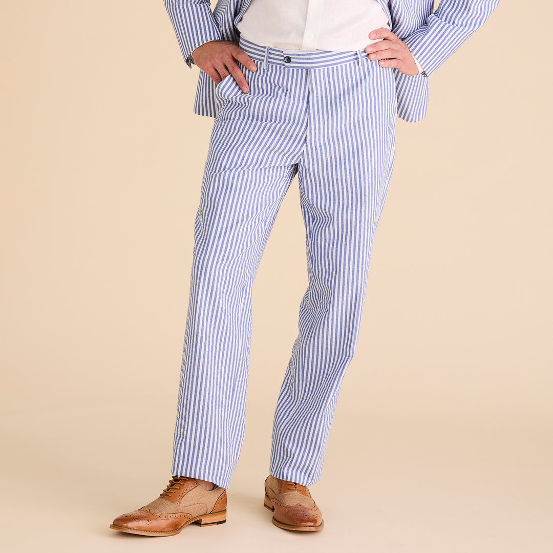 A person confidently stands with hands in pockets, dressed in Inserch Seersucker Stripe Pants P660155-00101 True Blue and a blue and white pinstripe suit with a modern fit, paired with a crisp white shirt and brown leather dress shoes.