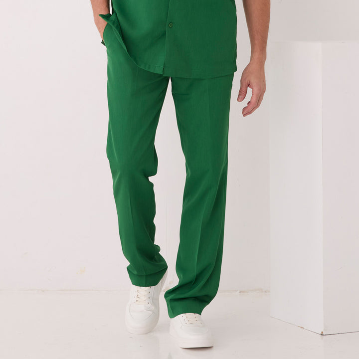 A person wears Inserch Crepe Flat Front Pants P9356-00200 Emerald with a matching green shirt, one hand in their pocket. White sneakers complete the look on a white floor with a white background.