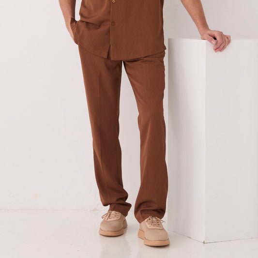 A person wears Inserch Crepe Flat Front Pants P9356-00064 Mocha, a matching brown shirt, and beige shoes, standing with one hand on a white block against a plain background.
