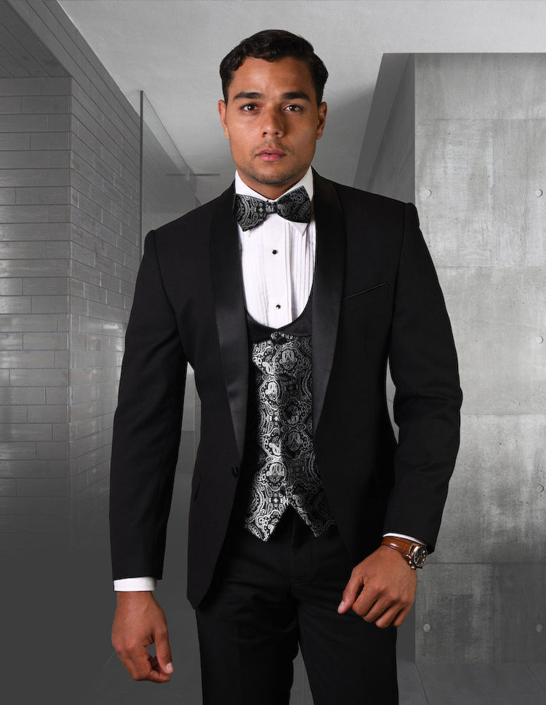A man in the STATEMENT CLOTHING | PALMERO-BLACK tuxedo by Statement Clothing, with a patterned black and white waistcoat and bow tie, stands in a chic, minimalist hallway.