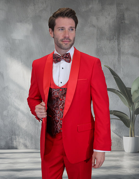 A man in the STATEMENT CLOTHING | PALMERO-RED tailored tuxedo by Statement Clothing, featuring a patterned vest and bow tie, stands indoors near a potted plant against a gray textured wall.