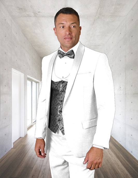A man in the STATEMENT CLOTHING | PALMERO-WHITE tuxedo by Statement Clothing stands in a modern minimalist hallway, showcasing flat front pants, a white suit jacket, and a patterned black vest with bow tie.