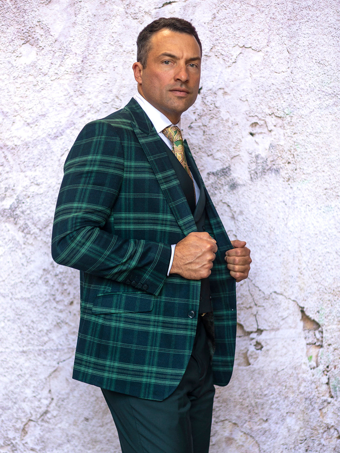 Dressed in the INSOMNIA PARIS-1 3PC Compose Tailored Fit Forest Suit by Insomnia, a man stands confidently before a textured, off-white wall, subtly holding the lapels of his jacket.