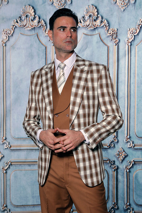 A man wears the Manzini PARIS-4 Men's 3-Piece Tailored-Fit Plaid Business Suit in Coco, featuring a plaid blazer and brown vest, standing before an ornate blue and white paneled wall.