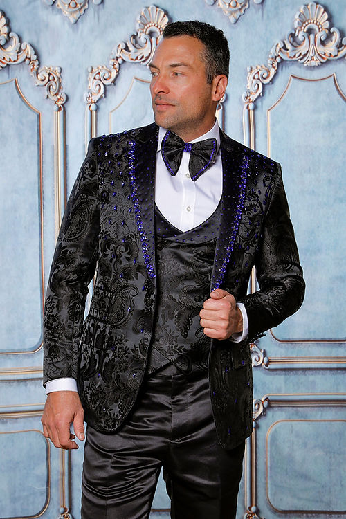 A man in an Insomnia MANZINI PLATINUM 3-Piece Slim Fit Rhinestone Satin Floral Black/Royal Tuxedo Suit, featuring a matching bow tie and vest, stands against an ornate blue wall.