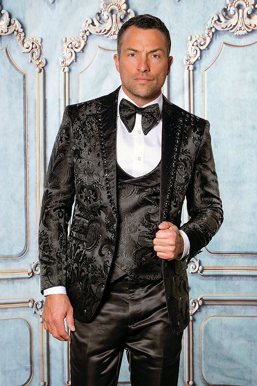 A man dons an Insomnia MANZINI PLATINUM 3-Piece Slim Fit Rhinestone Satin Floral Black Tuxedo Suit, with a bow tie, standing against an ornate blue and white backdrop, subtly catching the light with shimmering rhinestones woven into the fabric.