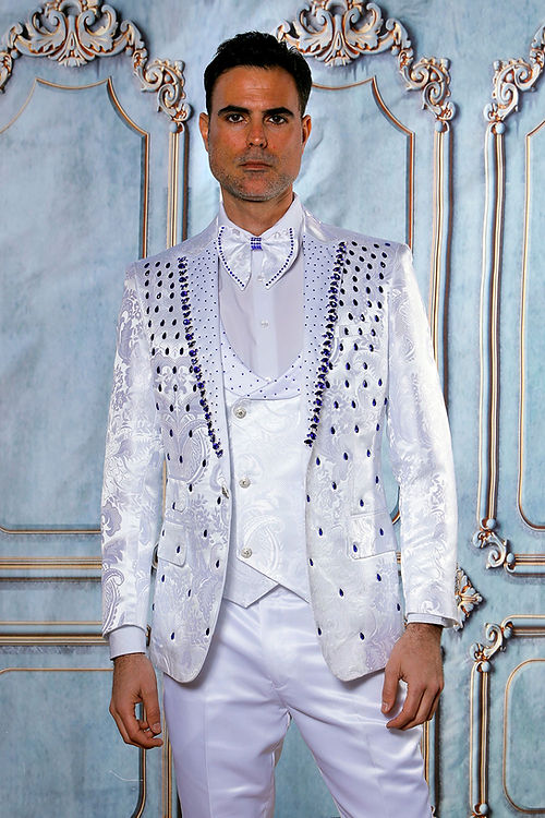 A person in the Insomnia MANZINI PLATINUM 3-Piece Slim Fit Rhinestone Satin Floral White/Royal Tuxedo Suit, complete with a bow tie, poses before an elaborate backdrop.