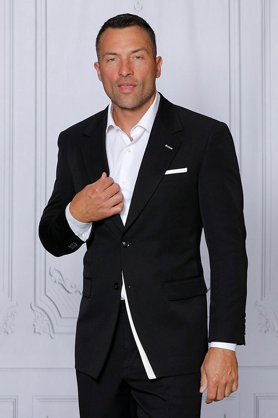 A man wears the INSOMNIA PURPLE LABEL 2PC Tailored Fit Black White 4 Way Stretch Spandex Suit by Insomnia with a white dress shirt, standing before a decorative white wall, looking at the camera and touching his jacket.