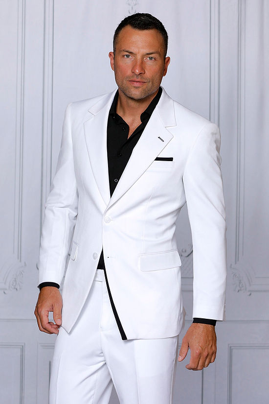 A man wears the INSOMNIA PURPLE LABEL 2PC Tailored Fit White Black 4 Way Stretch Spandex Suit by Insomnia with a black dress shirt, standing before a light paneled wall and looking at the camera.