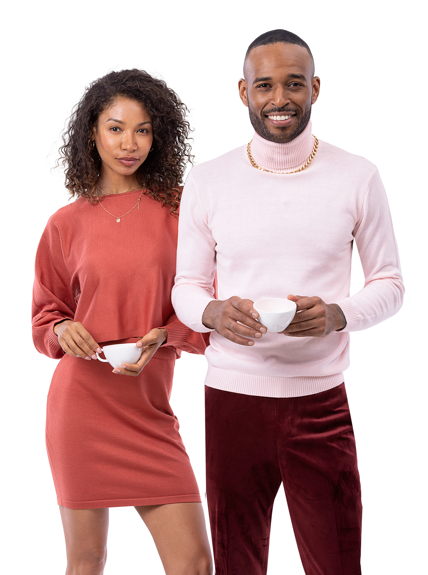 A man and a woman stand side by side holding white cups. Both are dressed in casual attire; the woman is wearing a red dress and the man is in a light pink sweater paired with EJ Samuel Wine Pants PV01.