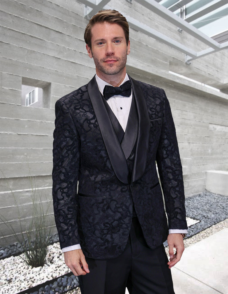 A man in the STATEMENT CLOTHING | REGENCY-BLACK tuxedo by Statement Clothing, with a white dress shirt and black bow tie, stands in a sleek outdoor space with concrete walls and stone landscaping.