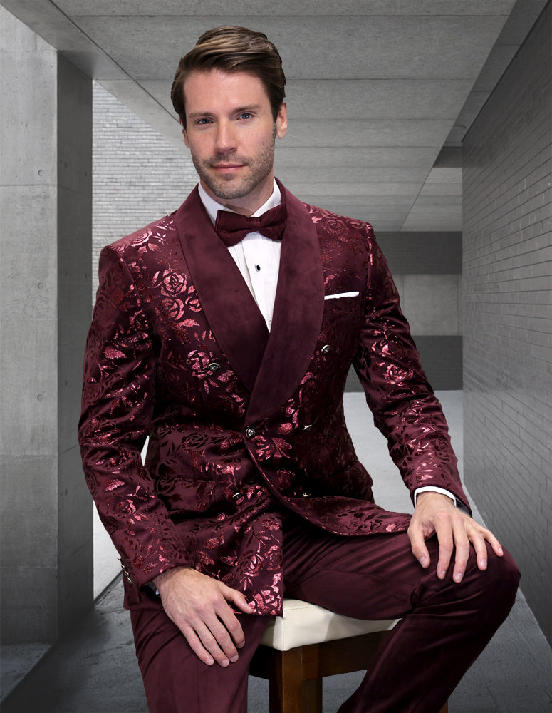 A man wearing the STATEMENT CLOTHING | RENESANSE-BURGUNDY tuxedo by Statement Clothing, paired with a bow tie, sits on a stool in a modern gray concrete corridor.