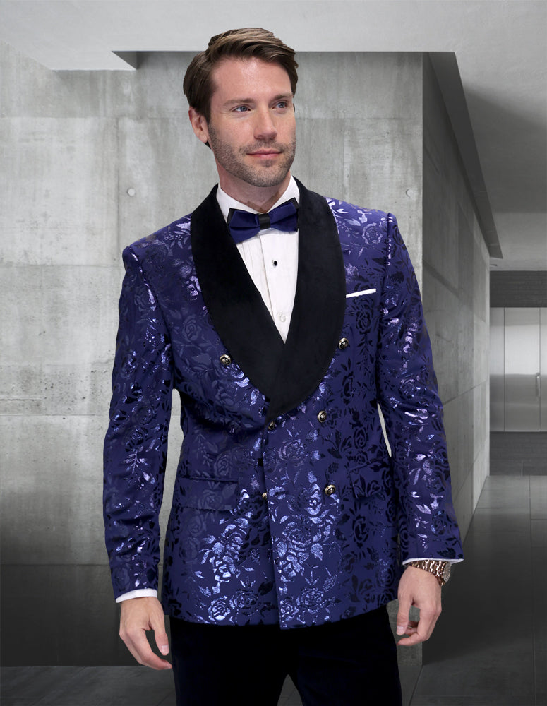 A man in the STATEMENT CLOTHING | RENESANSE-SAPPHIRE tuxedo by Statement Clothing—featuring a shiny blue floral pattern, black shawl lapel, white shirt, and black bow tie—stands in a modern, minimalist interior.