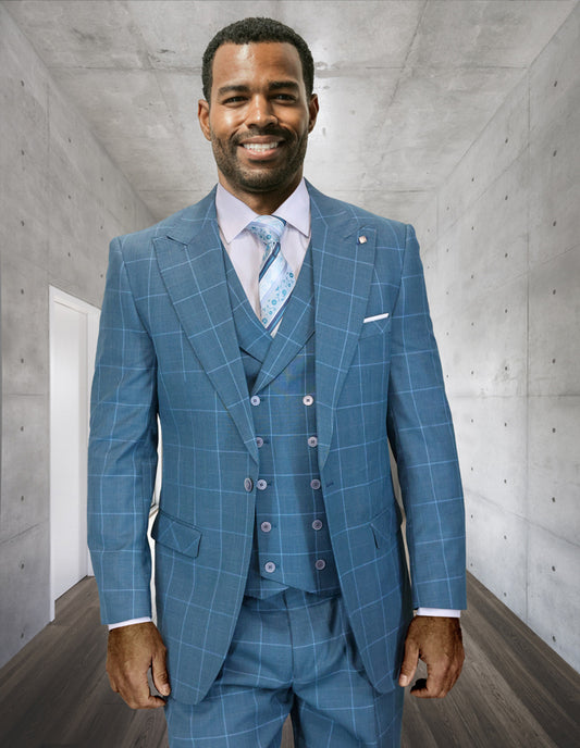 A man in the STATEMENT CLOTHING | RIGATO-JADE blue plaid suit by Statement Clothing, with a double-breasted vest and tie, smiles while standing in a modern hallway featuring concrete walls and a dark wood floor.