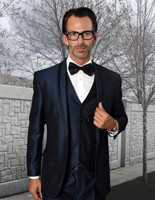 A man in the STATEMENT CLOTHING | RIO-INDIGO tailored suit by Statement Clothing, with a white shirt, black bow tie, and glasses, stands outdoors before a stone wall and leafless trees.