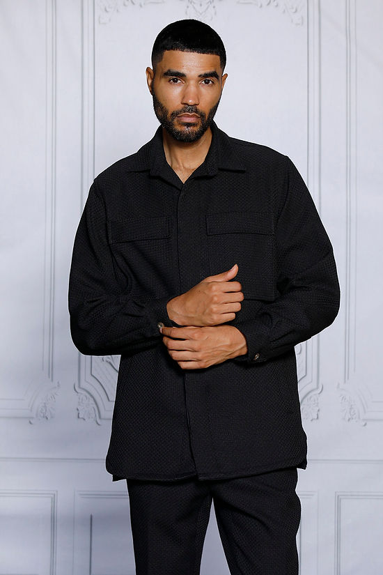 A man with short hair and a beard stands against a white decorative wall, wearing the Insomnia INSOMNIA ROBERTO 2PC Black Leisure Suit made from a soft poly rayon blend.