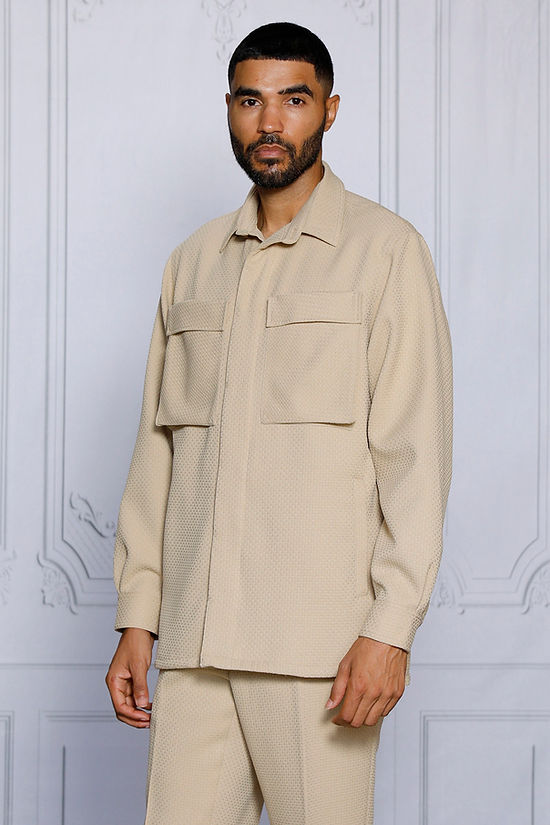 A man stands indoors in the INSOMNIA ROBERTO 2PC Tan Leisure Suit by Insomnia, featuring a textured button-up shirt with two large chest pockets and matching pants for a relaxed, casual look.