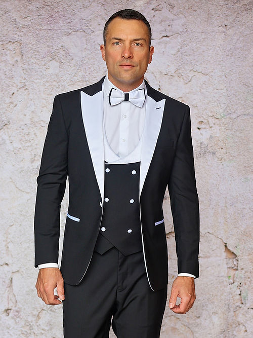 A man stands against a light-colored wall, wearing the Insomnia MANZINI ROME 3-Piece Slim Fit Stretch Spandex Black/White Tuxedo Suit with white lapels, a crisp white shirt, and a black bow tie.