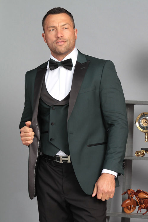 A confident man wearing the Insomnia MANZINI ROME Slim Fit 3-Piece Stretch Spandex Hunter Tuxedo Suit with black lapels and a bow tie stands with one hand on his jacket, accompanied by a vintage clock and sculpture in the background, enhancing the elegance of this dry clean only attire.