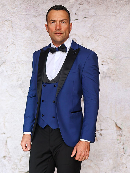 A man in an Insomnia MANZINI ROME Slim Fit 3-Piece Stretch Spandex Sapphire Tuxedo Suit with a black lapel and bow tie stands before a textured light wall, exuding elegance that requires dry clean only care.