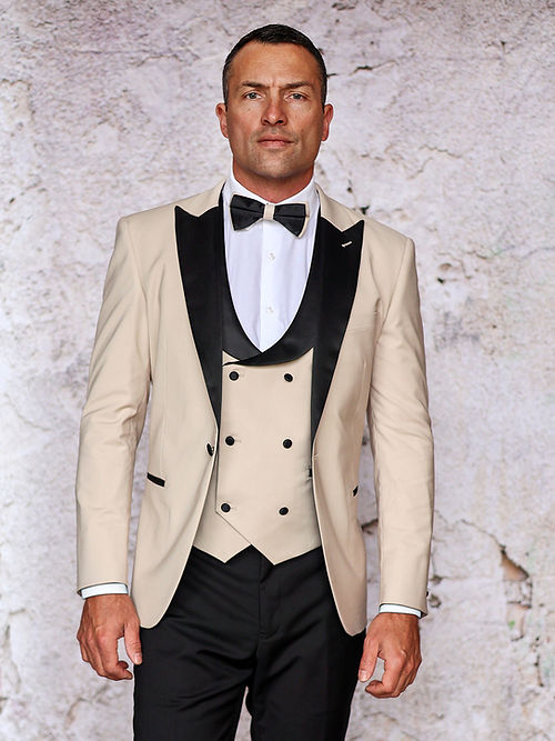 A man is wearing a black and beige MANZINI ROME Slim Fit 3-Piece Stretch Spandex Tan Tuxedo Suit by Insomnia, complete with a bow tie, standing against a textured wall.