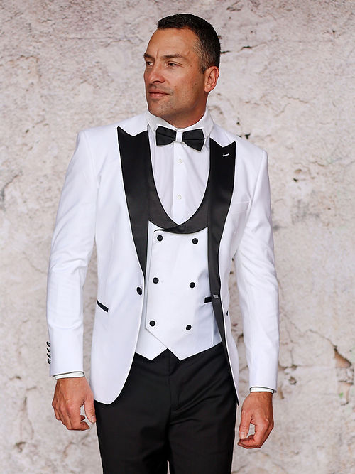 A man in the Insomnia MANZINI ROME 3-Piece Slim Fit Stretch Spandex White Tuxedo Suit stands before a textured light wall, featuring a white tuxedo jacket with black lapels, white waistcoat with black buttons, bow tie, and black pants.