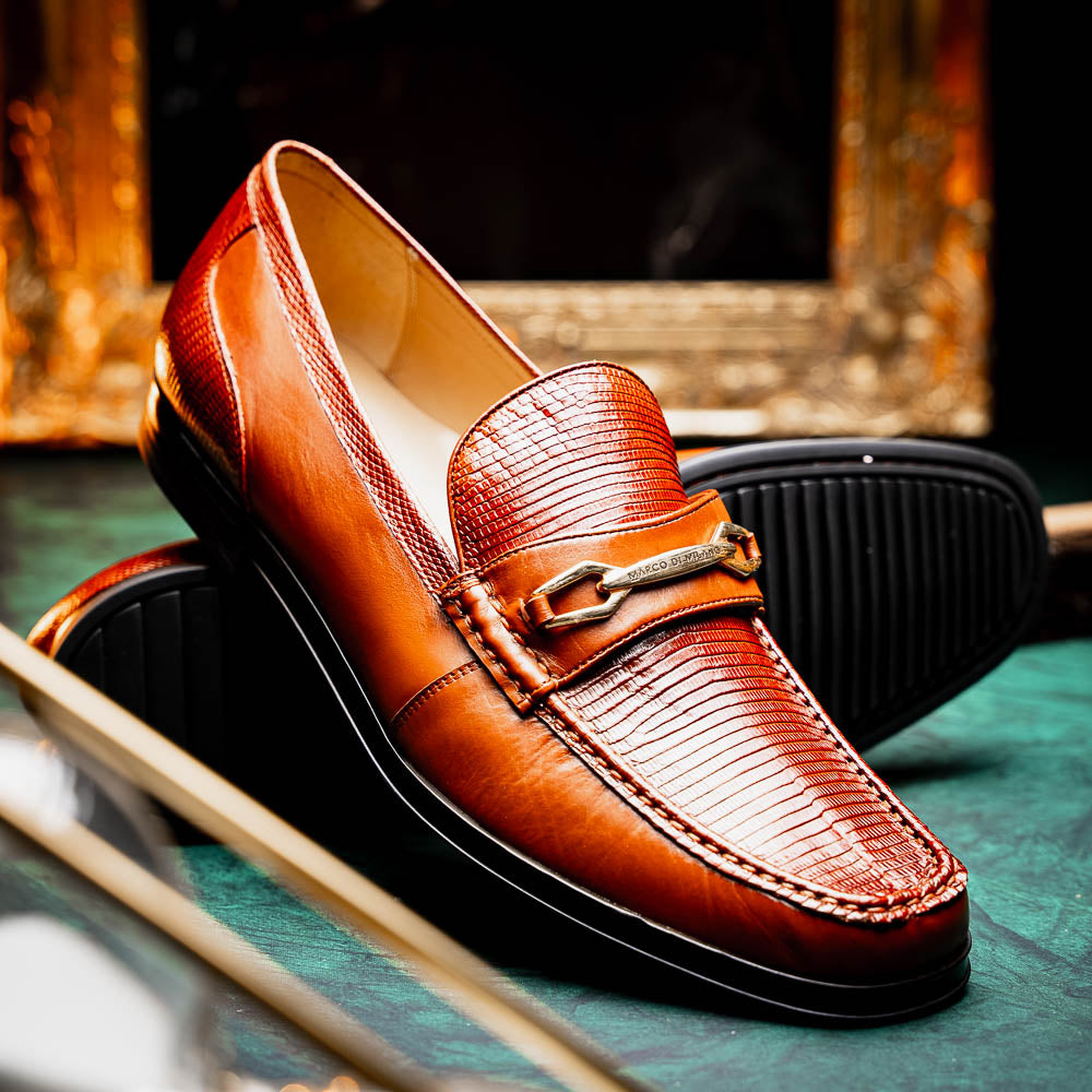The Marco Di Milano Ravello Cognac loafers feature brown leather with hand-painted finishes and metal detailing; one shoe is upright, while the other displays the black ridged sole.