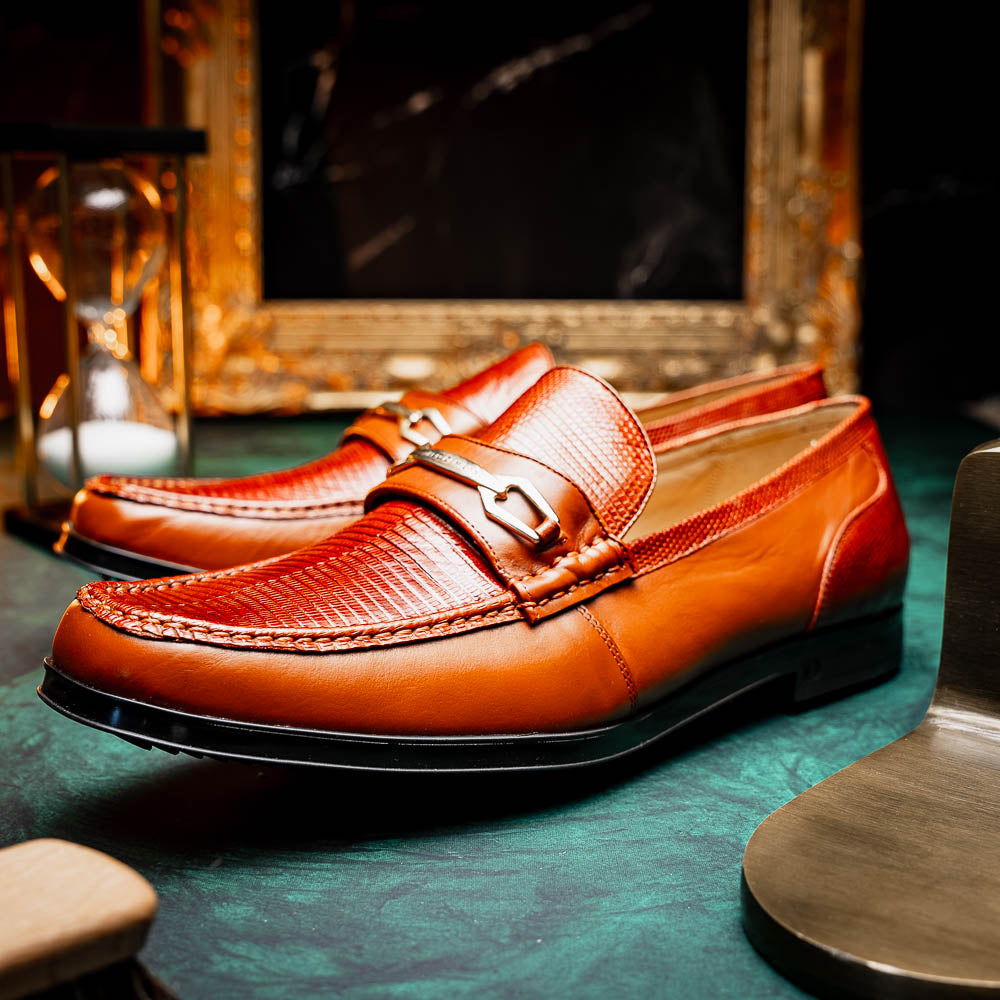 The Marco Di Milano Ravello Cognac loafers feature brown leather with hand-painted finishes and metal detailing; one shoe is upright, while the other displays the black ridged sole.