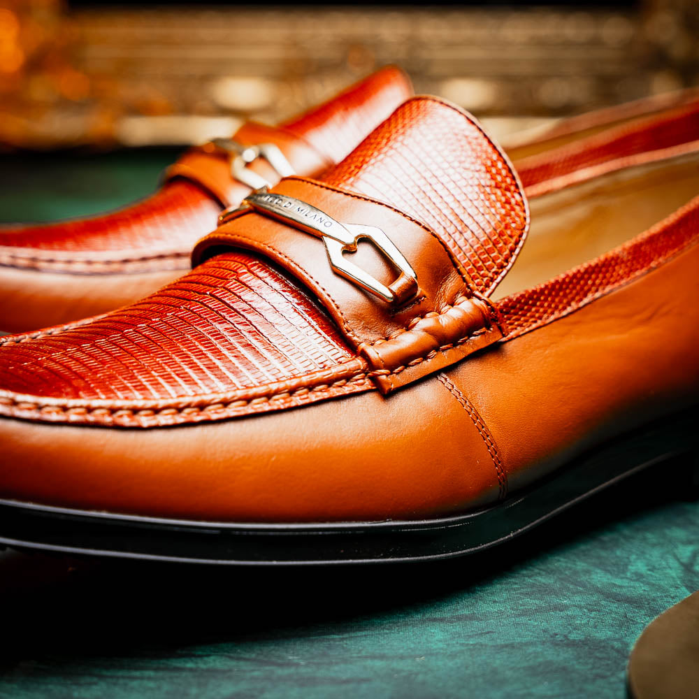 The Marco Di Milano Ravello Cognac loafers feature brown leather with hand-painted finishes and metal detailing; one shoe is upright, while the other displays the black ridged sole.