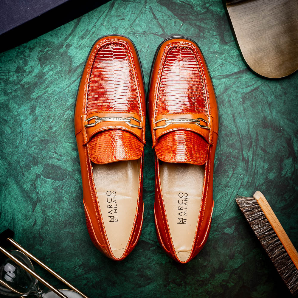 The Marco Di Milano Ravello Cognac loafers feature brown leather with hand-painted finishes and metal detailing; one shoe is upright, while the other displays the black ridged sole.
