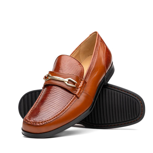 The Marco Di Milano Ravello Cognac loafers feature brown leather with hand-painted finishes and metal detailing; one shoe is upright, while the other displays the black ridged sole.