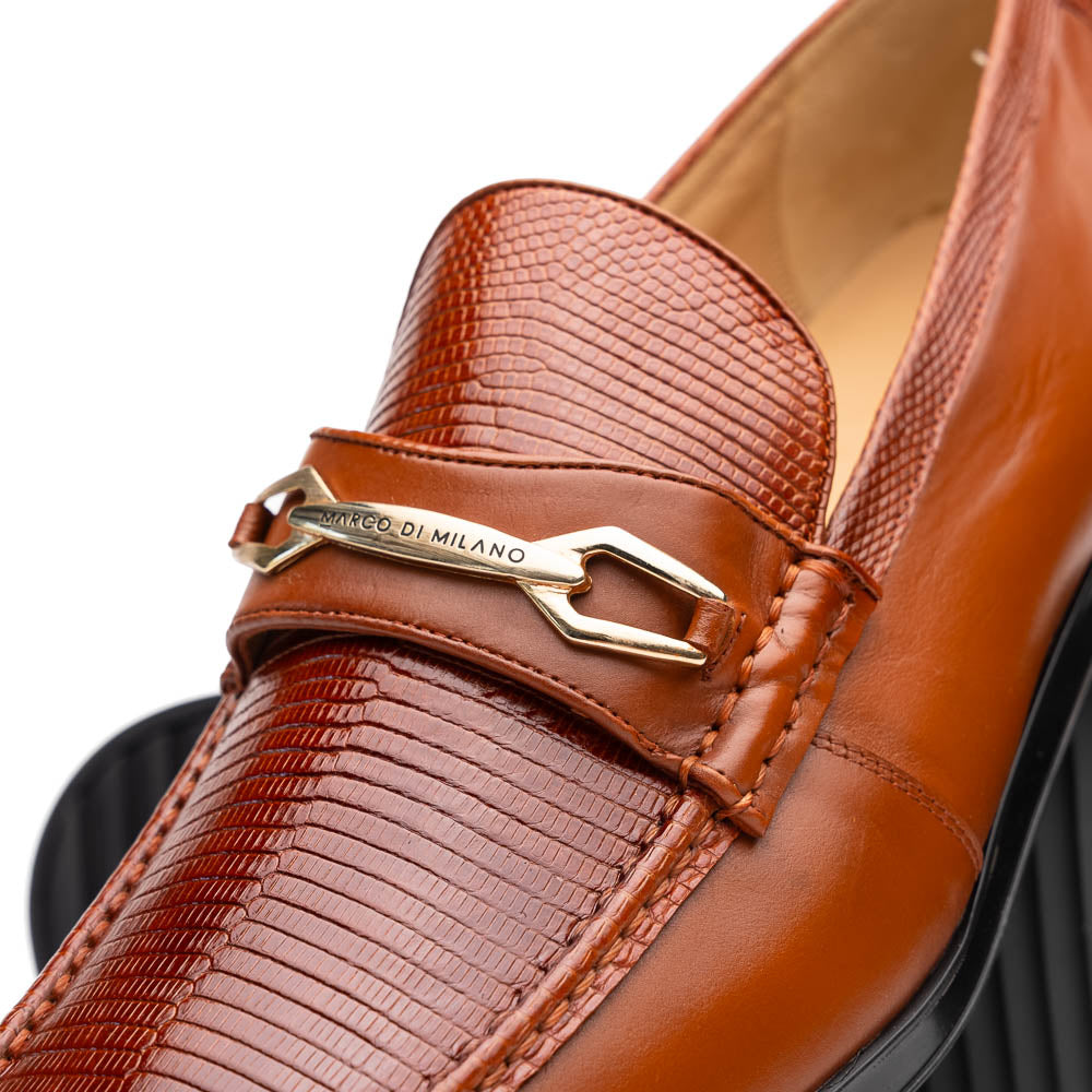 The Marco Di Milano Ravello Cognac loafers feature brown leather with hand-painted finishes and metal detailing; one shoe is upright, while the other displays the black ridged sole.