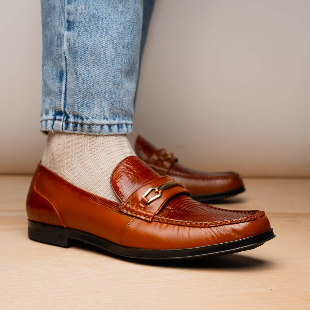 The Marco Di Milano Ravello Cognac loafers feature brown leather with hand-painted finishes and metal detailing; one shoe is upright, while the other displays the black ridged sole.