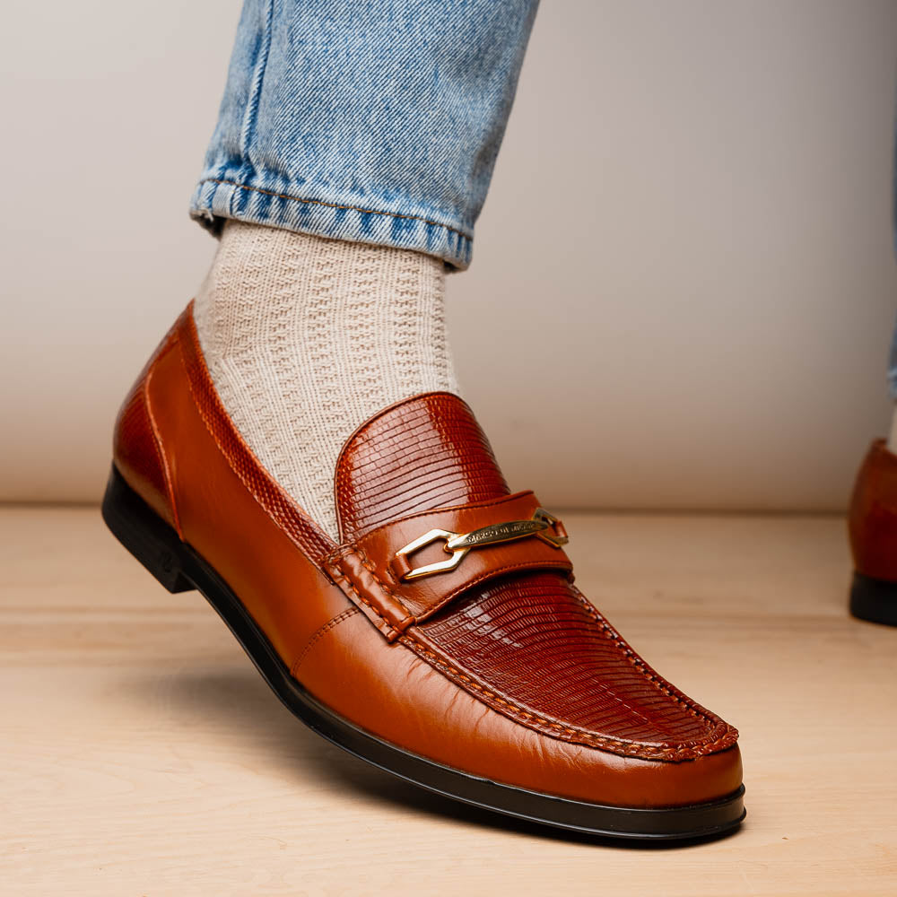 The Marco Di Milano Ravello Cognac loafers feature brown leather with hand-painted finishes and metal detailing; one shoe is upright, while the other displays the black ridged sole.