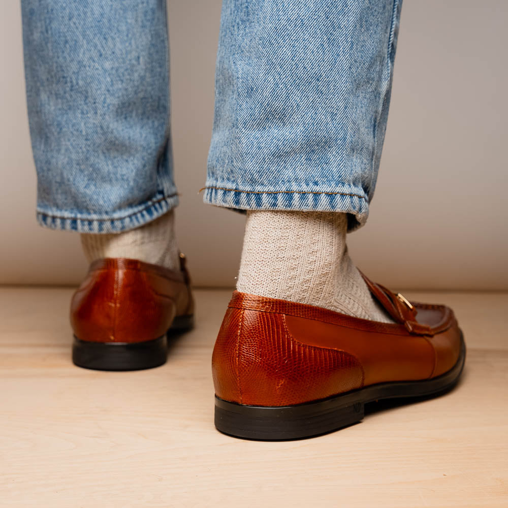 The Marco Di Milano Ravello Cognac loafers feature brown leather with hand-painted finishes and metal detailing; one shoe is upright, while the other displays the black ridged sole.