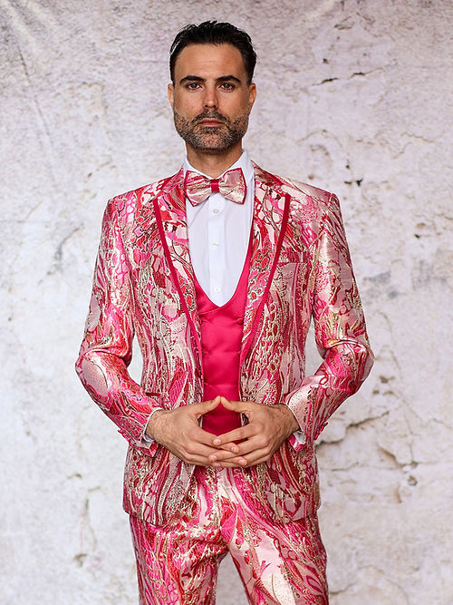 A man confidently stands against a textured light background in an Insomnia Manzini Santo 3-piece slim fit fuchsia tuxedo suit with a bright pink and gold metallic pattern, complemented by a matching bow tie.