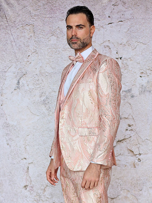 A man in an Insomnia MANZINI SANTO 3-piece slim fit pink tuxedo suit with a fancy metallic jacquard pattern stands against a textured white wall.
