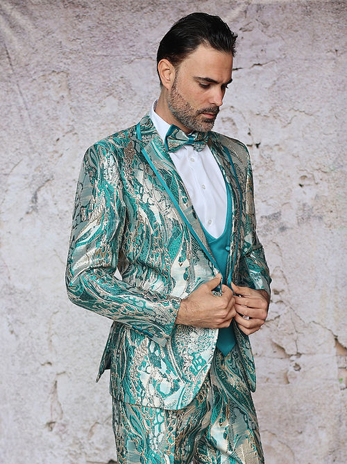 A man poses against a textured background in an Insomnia MANZINI SANTO turquoise tuxedo suit, featuring a fancy metallic pattern and bow tie, looking down.