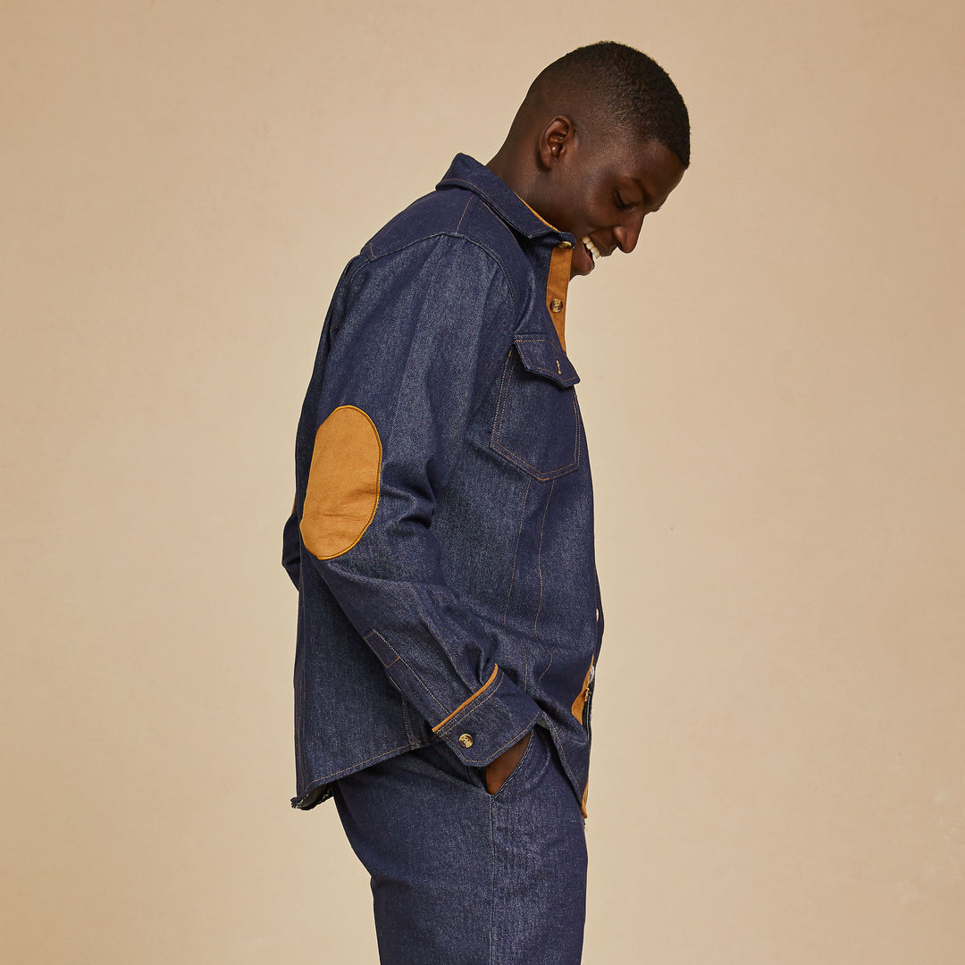 A person wears the Inserch Denim Two-Pocket Shirt SE003A-10 in Denim Blue by Inserch, paired with a blue and white houndstooth sweater and matching denim pants, standing against a beige background.