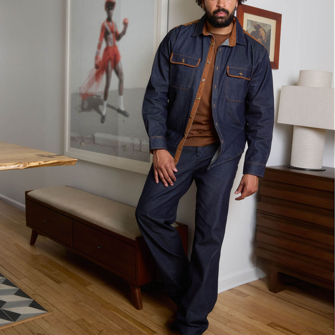 A man stands against a beige background wearing the Inserch Denim Shirt w/ Suede Trim 2pc Set in indigo by Inserch, paired with an orange sweater and dark brown shoes.