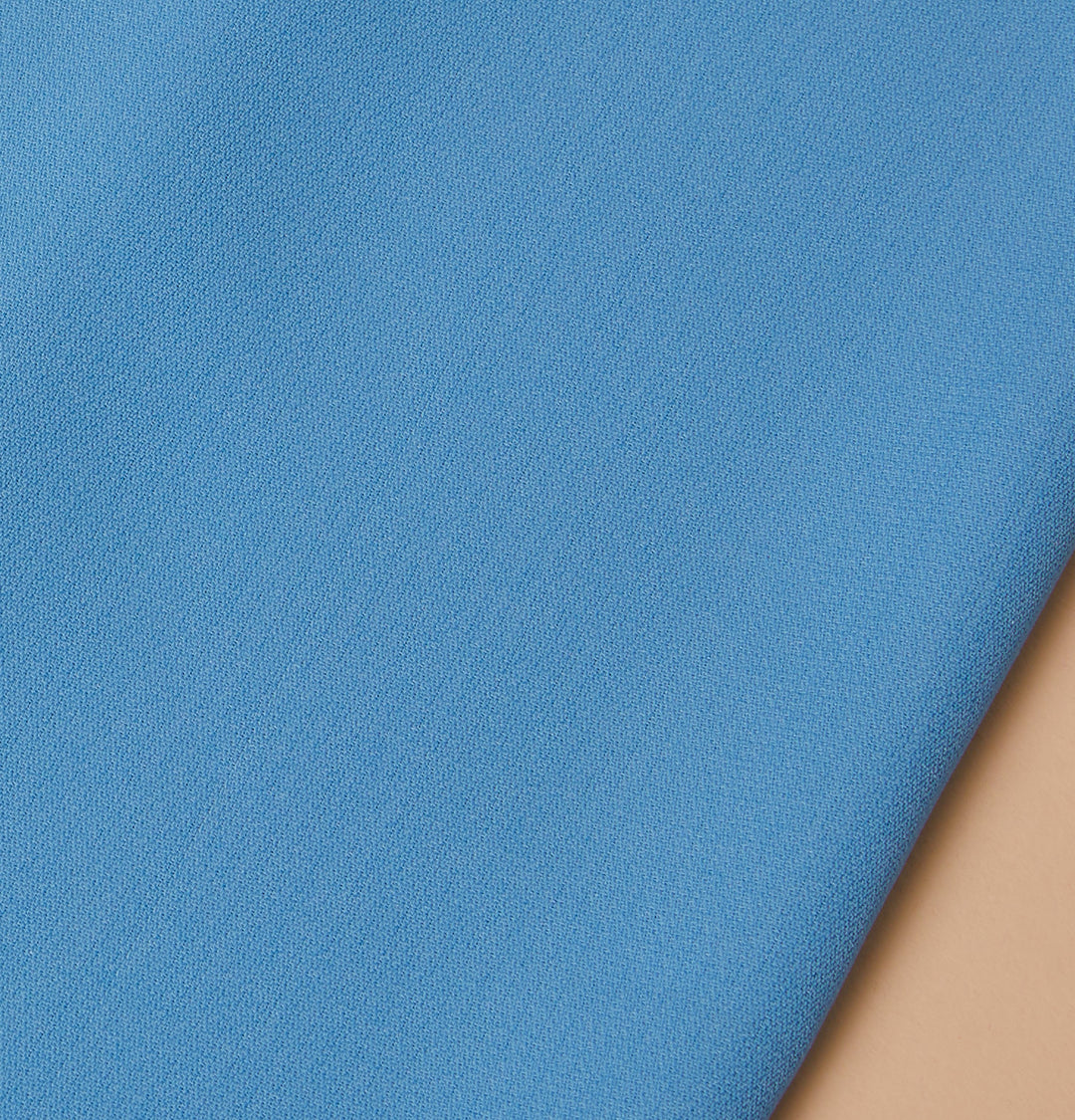 A close-up of Inserch Crepe Camp Collar Shirt fabric in Blue Mist, made with high-twist poly-crepe, ideal for a regular fit, displayed on a beige surface. Available as style SE9356 in Plum, Summer Pink, and Blue Mist.