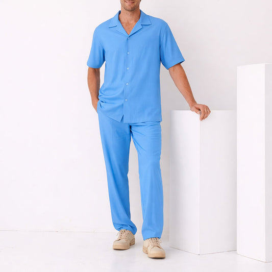 A man stands indoors wearing the Inserch Crepe Camp Collar Shirt SE9356-00183 Blue Mist with matching pants and beige sneakers, resting one hand on a white rectangular pedestal.