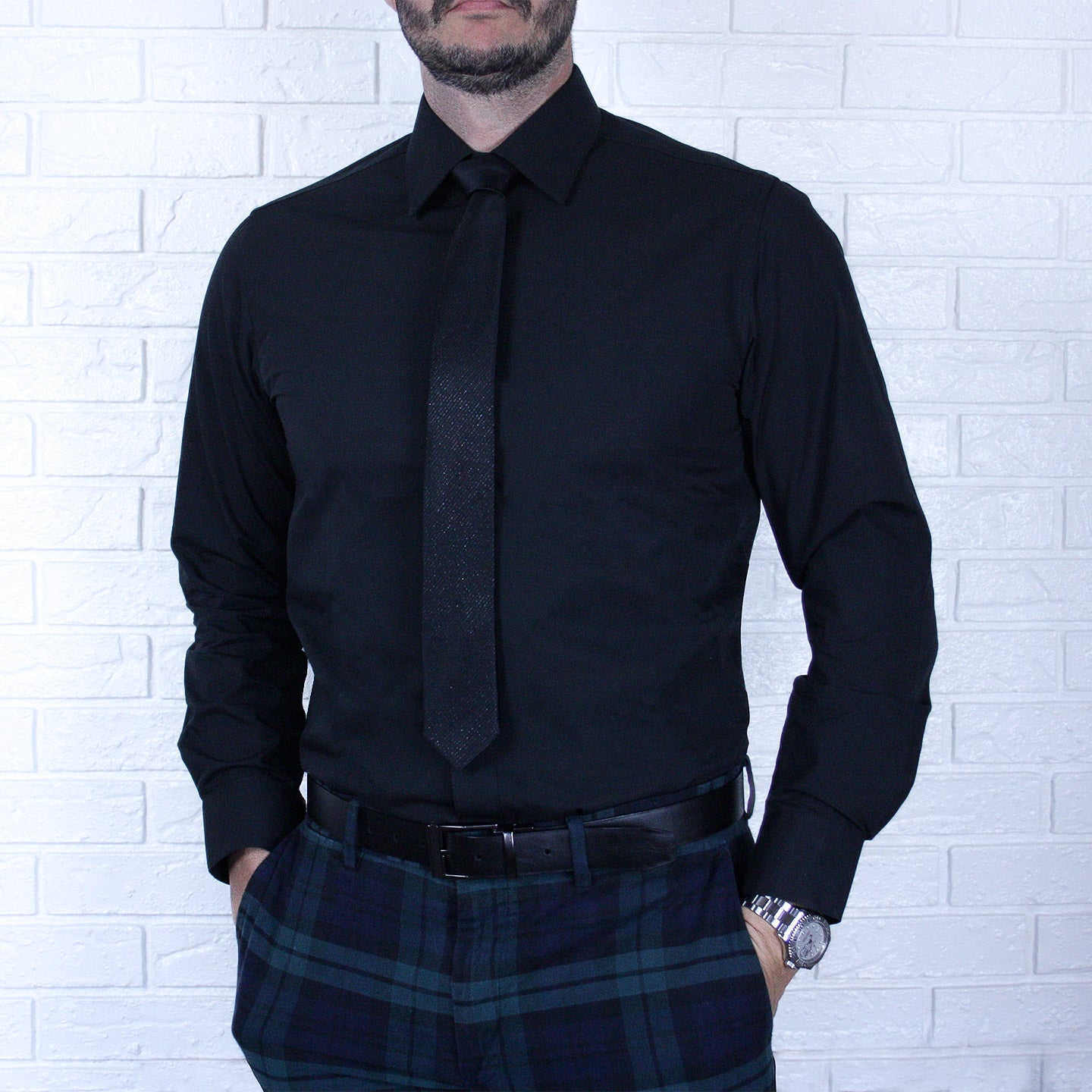 A man wearing an ARTURO Slim Fit Long Sleeve Black Dress Shirt, a black tie, and plaid pants stands against a white brick wall.