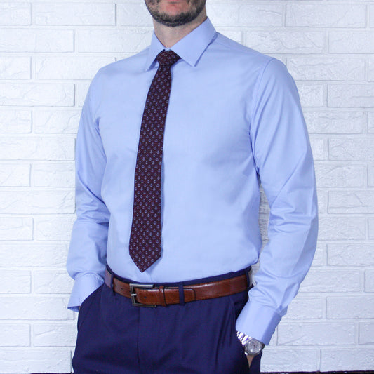 A man in an ARTURO Slim Fit Long Sleeve Light Blue Dress Shirt with a patterned tie, brown belt, and dark polyester pants stands against a white brick wall.